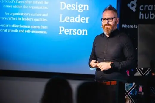 Klein Péter mikroportos előadás közben, mögötte kék dián a Design Leader Person felirat és három angol nyelvű tétel.